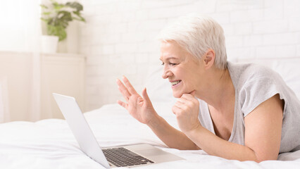 Positive elderly woman is reclining on a bed with a laptop computer open in front of her, waving at computer screen, have video call