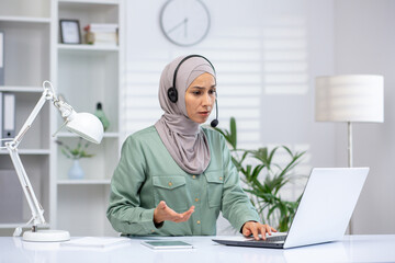 Businesswoman wearing a headset and hijab, working on a laptop during a virtual meeting in a modern office. Professional, focused, and engaged in an online discussion.