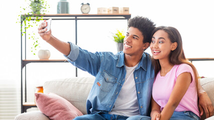 A teen couple sits on a couch in a living room and takes a selfie with a smartphone. The guy is...