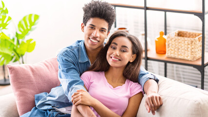 Black teen couple relaxing together on a couch in a living room. The guy has his arm around the girl, and they are both smiling and looking at the camera.