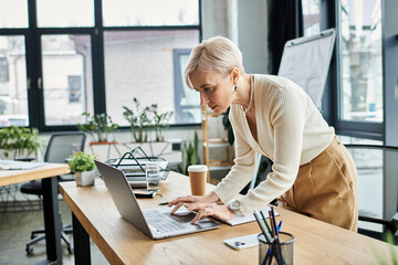 Middle-aged businesswoman with short hair focused on her laptop in a modern office setting.