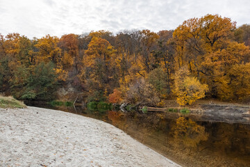 Autumn golden nature by sandy river shore with cloudy sky and mirror reflection on calm water surface