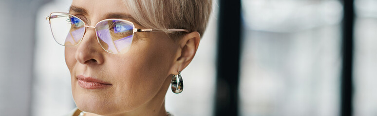 A middle-aged businesswoman with short hair wearing glasses gazes thoughtfully to the side in a modern office setting.