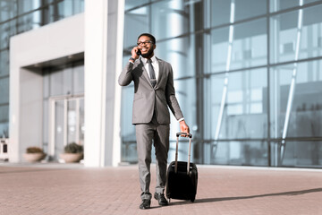 An African American businessman with baggage on a call at the airport entrance. The scene is busy and modern, highlighting the professional's travel routine.