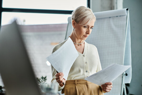 A middle-aged businesswoman with short hair holds a piece of paper in front of a computer screen.