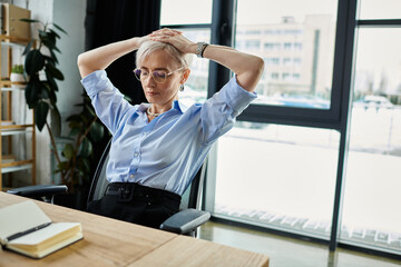 Middle-aged businesswoman sits at desk with hands on head, deep in thought. Short hair and...
