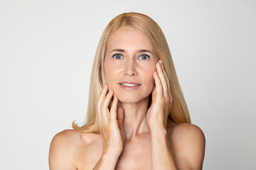 A close-up portrait of a middle-aged blonde woman with blue eyes, touching her face with both hands. She is looking at the camera with a slight smile. Her skin is smooth and flawless.