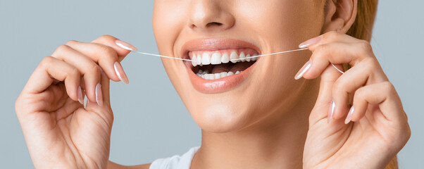 A close-up shot of a woman smiling while flossing her teeth with white floss against a grey...