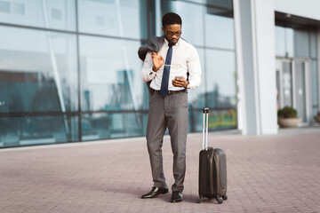 African American businessman is making a call by the entrance of a modern airport, with luggage beside him. The mood is professional and active, showcasing travel and communication.