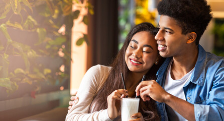 African American young guy and girl are seated at a table in a cafe, both holding milkshakes and sipping through straws. They appear relaxed and engaged in conversation, copy space