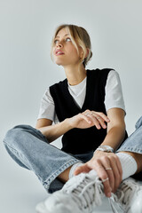 Fototapeta premium A young woman with blonde hair sits gracefully on the floor, legs crossed, in stylish casual attire against a grey backdrop.