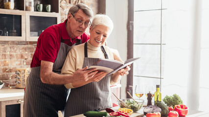 An older couple wearing aprons is reading cooking book in their kitchen. They are smiling and...