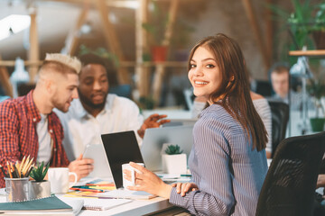 Group of young colleagues working together on a project, discussing ideas, and using laptops in a well-lit, modern office space.