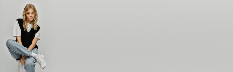 A stylish young woman with blonde hair elegantly poses on a stool against a grey backdrop.