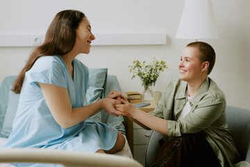 Pregnant patient sitting on bed and holding lovely her friends hands while they having lively talk