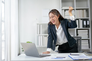 Excited businesswoman celebrating success at work while looking at laptop in modern office setting with documents on desk.