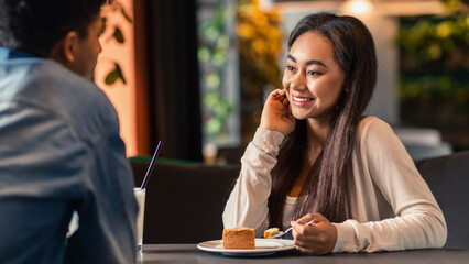 African American guy and girl are seated at a table in a bustling cafe. They are engaged in conversation, with coffee cups in front of them. The cafe is decorated with plants