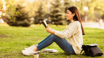 Asian woman is sitting on the grass, engrossed in a book she is reading. The sunlight highlights...