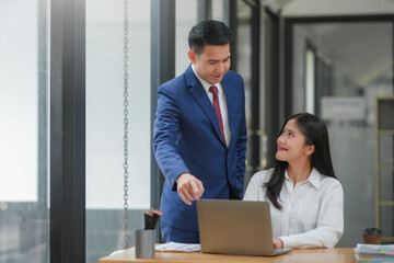 Professional business meeting with a man in a blue suit and a woman in white shirt discussing work in a modern office with a laptop.