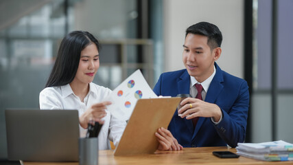 Two colleagues reviewing documents at the office, discussing business strategy and project ideas, with a laptop on the table.