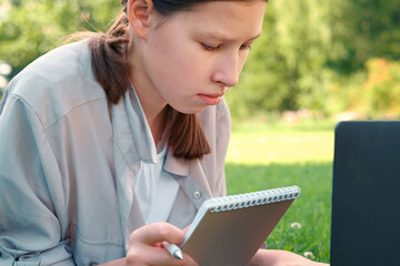 Fototapeta premium Teenage schoolgirl studying reading her books, tablet and notebook, sitting outdoors. Back to school. Student girl lying on the green grass using laptop in the college yard or park. Distance learning.