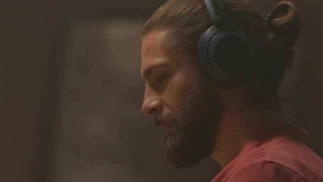 360 degree time lapse home. Close-up portrait of young handsome smiling Caucasian man with headphones listening to music. Guy working and enjoying song in evening. Living room. Camera moving around
