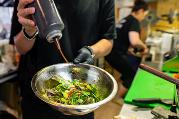 A chef in a commercial kitchen pours sauce onto a fresh salad.
