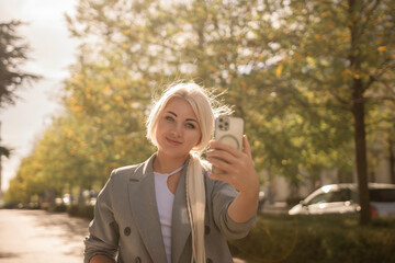 A woman is taking a picture of herself with her cell phone. She is wearing a gray jacket and scarf. The scene is set in a city with trees and cars in the background.