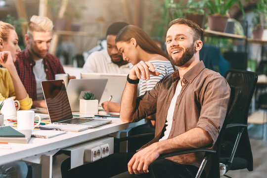 A smiling man sits at a desk in a modern office while his colleagues engage in a collaborative work session around him.