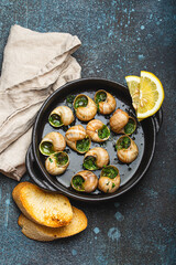 Escargots de Bourgogne Snails with Garlic Butter and Parsley in black cast iron pan with Lemon and Toasted Baguette Slices on rustic stone background top view, traditional French Delicacy .
