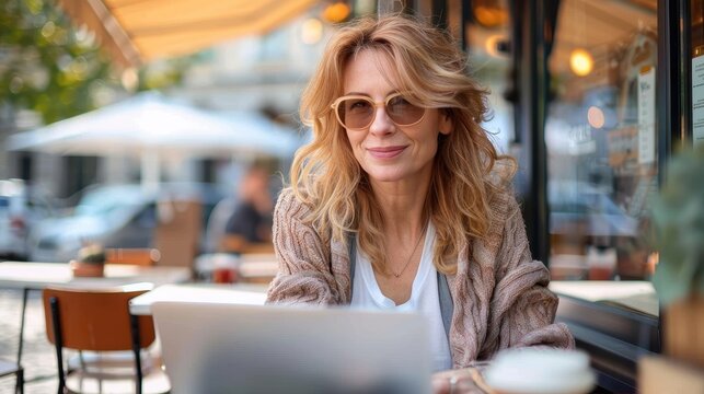 A joyful middle-aged woman wearing glasses and a cozy sweater is sitting at a cafe table with her laptop, possibly working or browsing