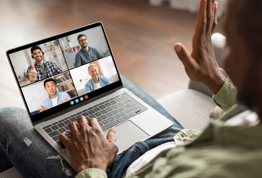 A man is sitting on a couch and participating in a video conference call with three other colleagues on his laptop. He is using his hand to gesture and is clearly engaged in the conversation
