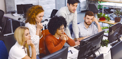 A group of focused multiethnic colleagues are seated at desks using desktop computers in a well-lit, contemporary office. They appear to be engrossed in work, displaying concentration and teamwork