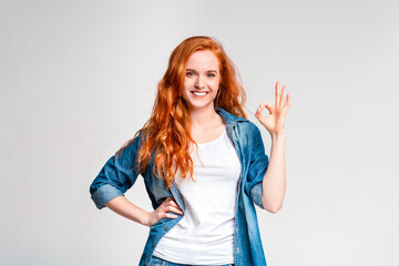 A cheerful woman with long red hair is standing confidently with one hand on her hip and the other...