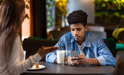African American young guy is seen attentively looking at his smartphone while sitting at a table in a cozy cafe. Another person, who is partially visible, appears to be talking to him.