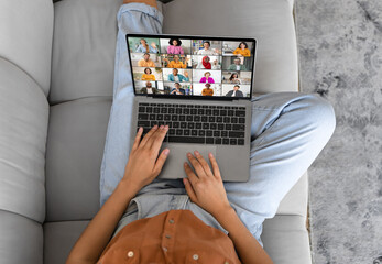 Unrecognizable lady is sitting on a couch and using a laptop to video call with several other people. The person is using the trackpad on the laptop to navigate the call.