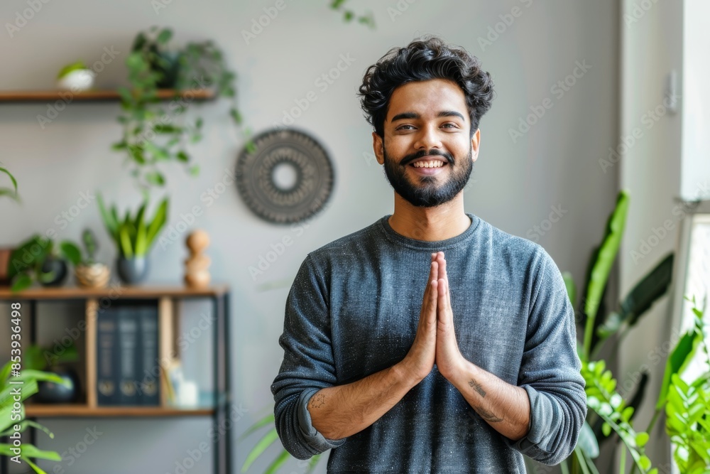 Wall mural Portrait of a cheerful indian man in his 20s joining palms in a gesture of gratitude isolated in modern minimalist interior - Wall murals