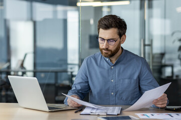 A serious and focused young man businessman sits in the office at a desk with a laptop, writes with...
