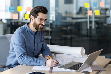 A young smiling male architect, engineer and developer works in the office according to drawings,...