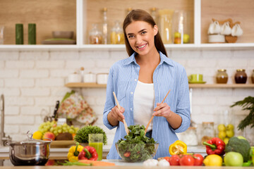A woman smiles while mixing a salad in her kitchen, surrounded by fresh vegetables and a pot on the counter.