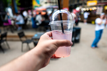 Close-up of a hand holding a pink drink with a straw in a plastic cup, against an unfocused crowd backdrop at an outdoor venue.