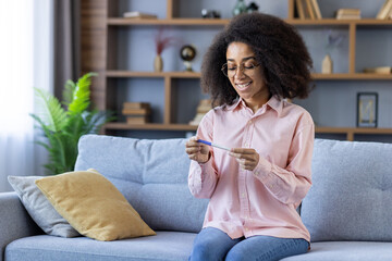 Obraz premium Happy woman holding a positive pregnancy test while sitting on a couch in a comfortable and cozy living room.