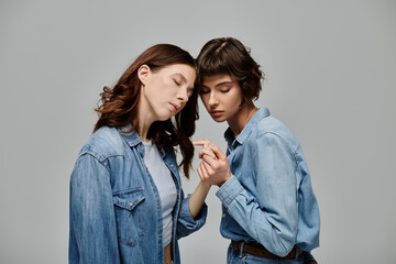 Two young women in denim attire pose intimately against a grey background, showcasing their love and style.