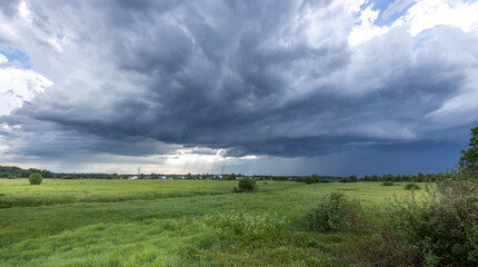 A dark, ominous storm cloud hangs low over a green field, threatening rain. The sun shines brightly through breaks in the clouds, illuminating the lush landscape.