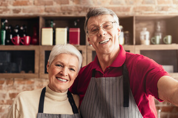 An elderly couple, wearing aprons, is filming a cooking show in their home kitchen. The man is in the foreground, gesturing towards the camera while smiling, taking selfie