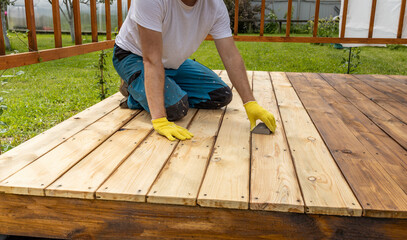 A man wearing yellow gloves sands a wooden deck in the backyard. The deck is made of light-colored...