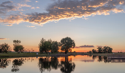 Naklejka premium A serene sunset paints the sky with hues of orange and pink, reflecting off the tranquil waters of a park lake. Trees line the shore, creating a picturesque backdrop for a peaceful evening.