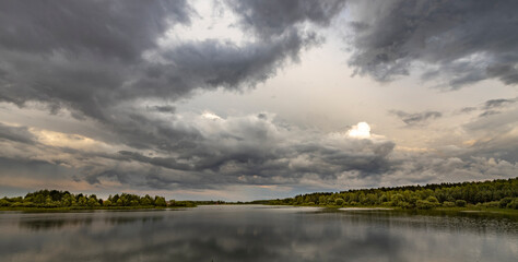 A peaceful lake mirrors a row of trees under stormy clouds. The setting sun adds a warm glow to the horizon, creating a beautiful contrast.
