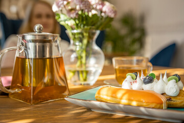 Teapot of tea, cream puffs with cream & blueberries on a table. Flowers in a vase & a womans silhouette in the background.