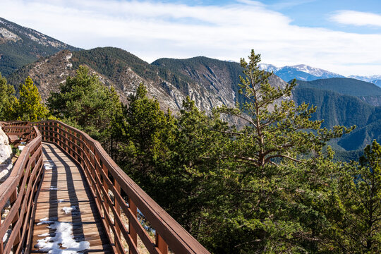 A scenic view with a wooden walkway leading through a mountainous landscape, with snow patches and green trees under a blue sky